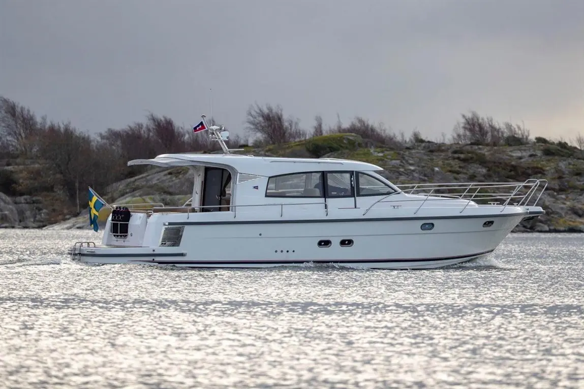 White motor yacht gliding across water, featuring Swedish flag, rocky shore in background, under overcast sky.